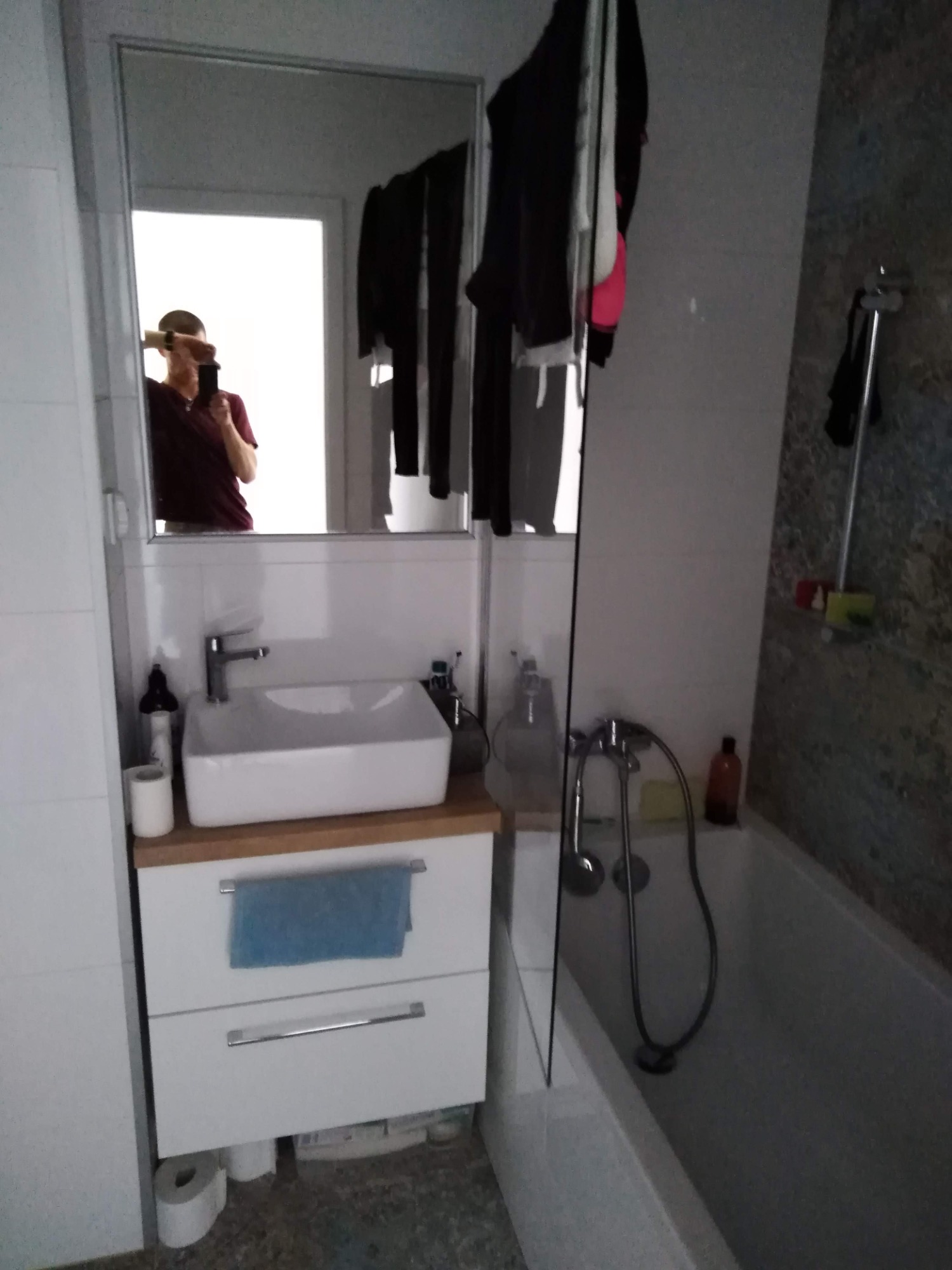 Nowoczesna łazienka z białą umywalką nablatową, szafką pod umywalką z niebieskim ręcznikiem, wanną i lustrem, w którym odbija się osoba robiąca zdjęcie.