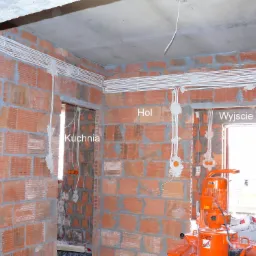 Rozprowadzone przewody elektryczne w surowym wnętrzu z cegły, widoczne puszki instalacyjne i pomarańczowa maszyna do zaprawy w tle.