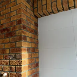 Wykończenie narożnika i łuku cegłą klinkierową w odcieniach czerwieni i brązu, z szarą fugą, przylegające do białych, gładkich płytek.