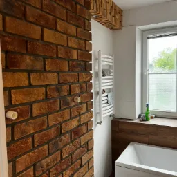 Ceglana ściana z widocznymi instalacjami w nowocześnie urządzonej łazience z białymi płytkami, grzejnikiem drabinkowym i oknem z matową folią.