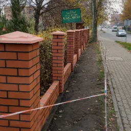 Ceglany murek z filarami i daszkami w kolorze terakoty, budowany wzdłuż chodnika i ulicy, z widocznym znakiem 'Gościniec Mazurski' w tle, otoczony taśmą ostrzegawczą.