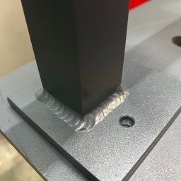 PROLAMIX JANUSZ LAMIK - Spoina łącząca czarny, kwadratowy profil z szarą, metalową płytą. Widoczne ślady spawania, otwór w płycie. Detal konstrukcji stalowej.