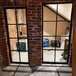 PROLAMIX JANUSZ LAMIK - Dwa okna z czarnymi szprosami w murze z czerwonej cegły. W tle widoczne przedmioty w pomieszczeniu, w tym skrzynki i torba. Widoczne cienie na podłodze.