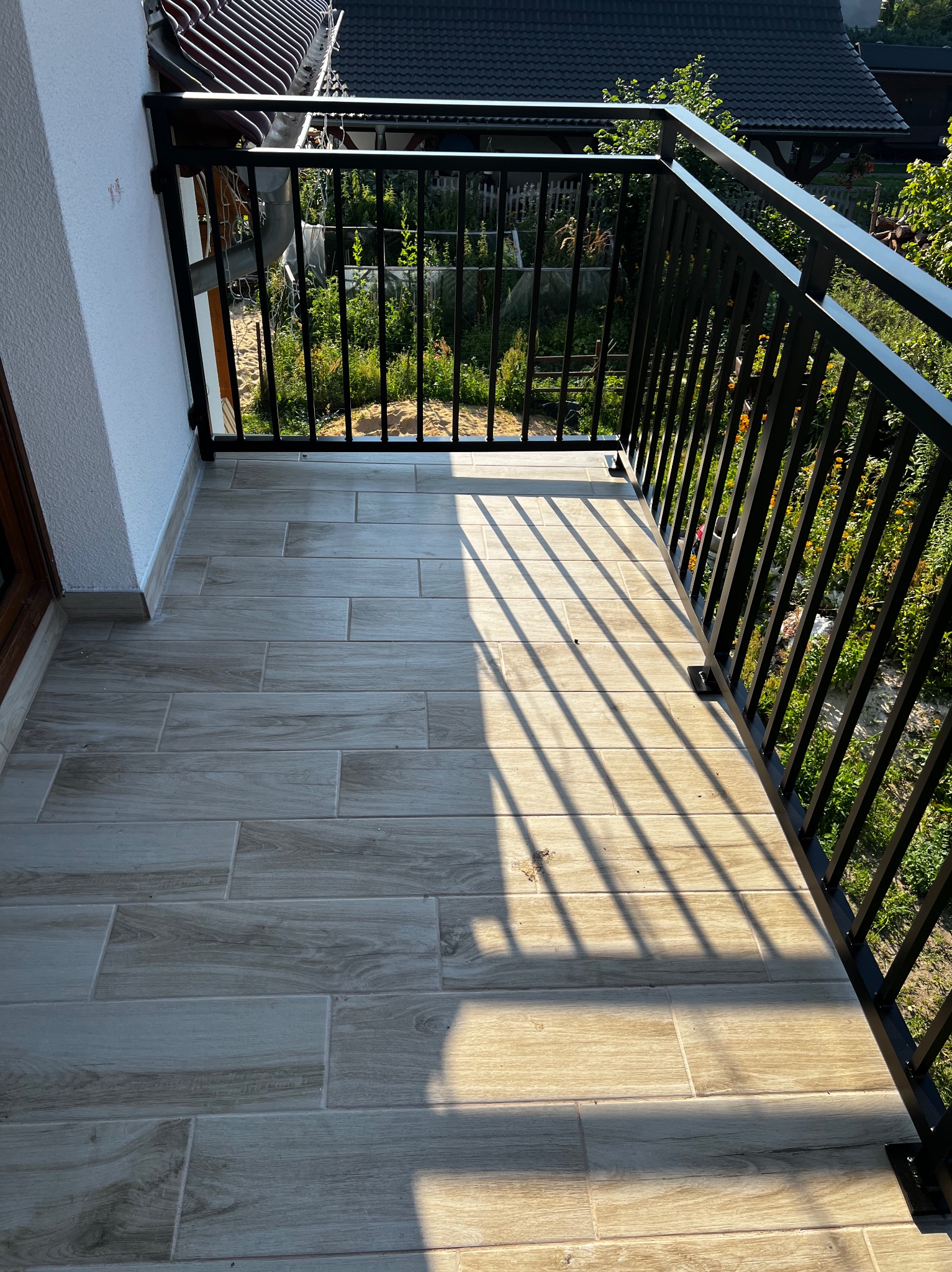 Czarna, metalowa balustrada balkonowa z pionowymi szczebelkami, z widokiem na ogród. Jasne płytki podłogowe na balkonie, oświetlone promieniami słońca.