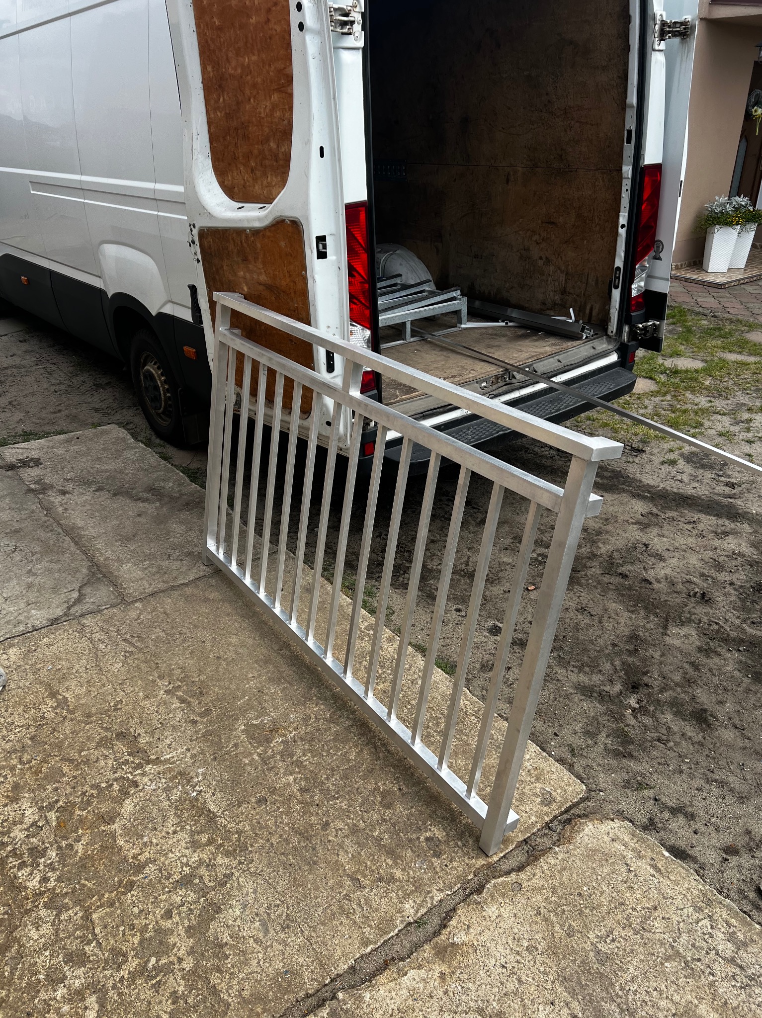 Część balustrady aluminiowej oparta o betonowy chodnik, przy otwartym samochodzie dostawczym. Widoczny fragment posesji z roślinami w tle.