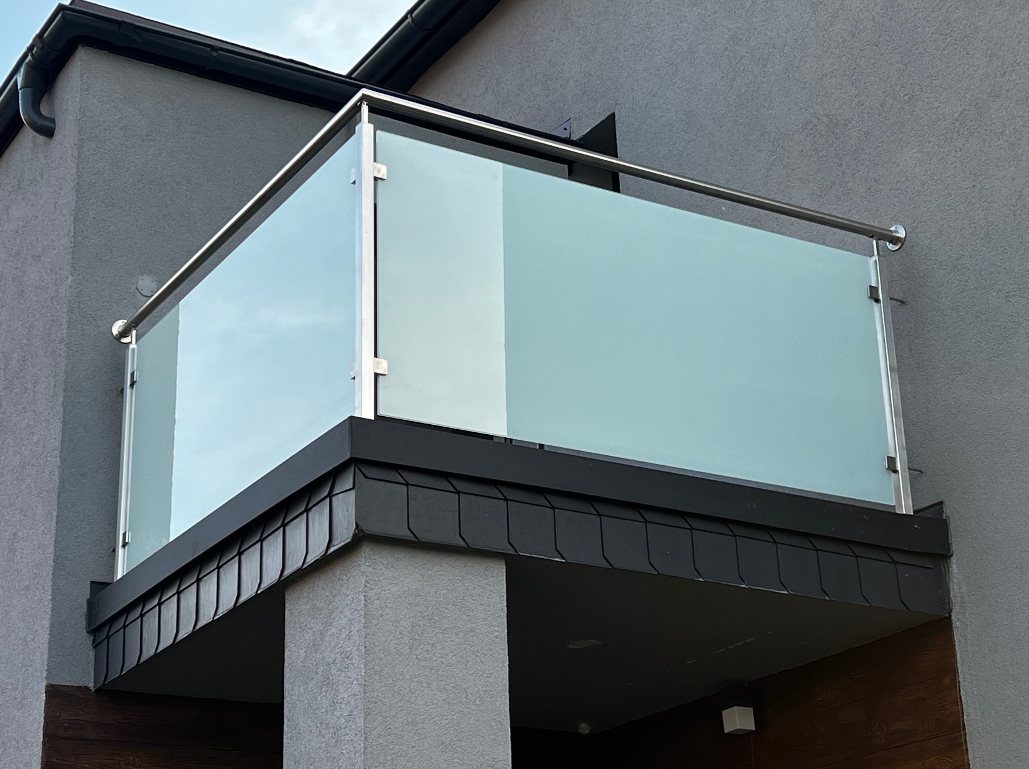 Nowoczesna balustrada balkonowa ze stali nierdzewnej i matowego szkła, zamontowana na szarym budynku z ciemnym dachem. Minimalistyczny design, eleganckie wykończenie.