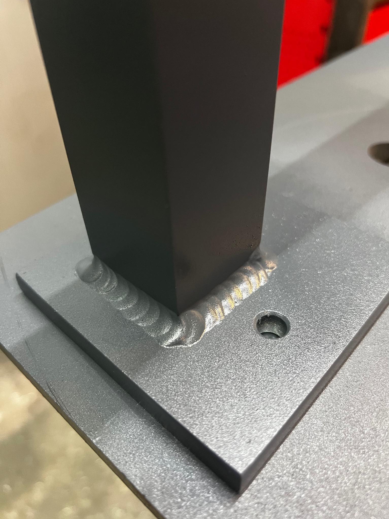 Spoina łącząca czarny, kwadratowy profil z szarą, metalową płytą. Widoczne ślady spawania, otwór w płycie. Detal konstrukcji stalowej.