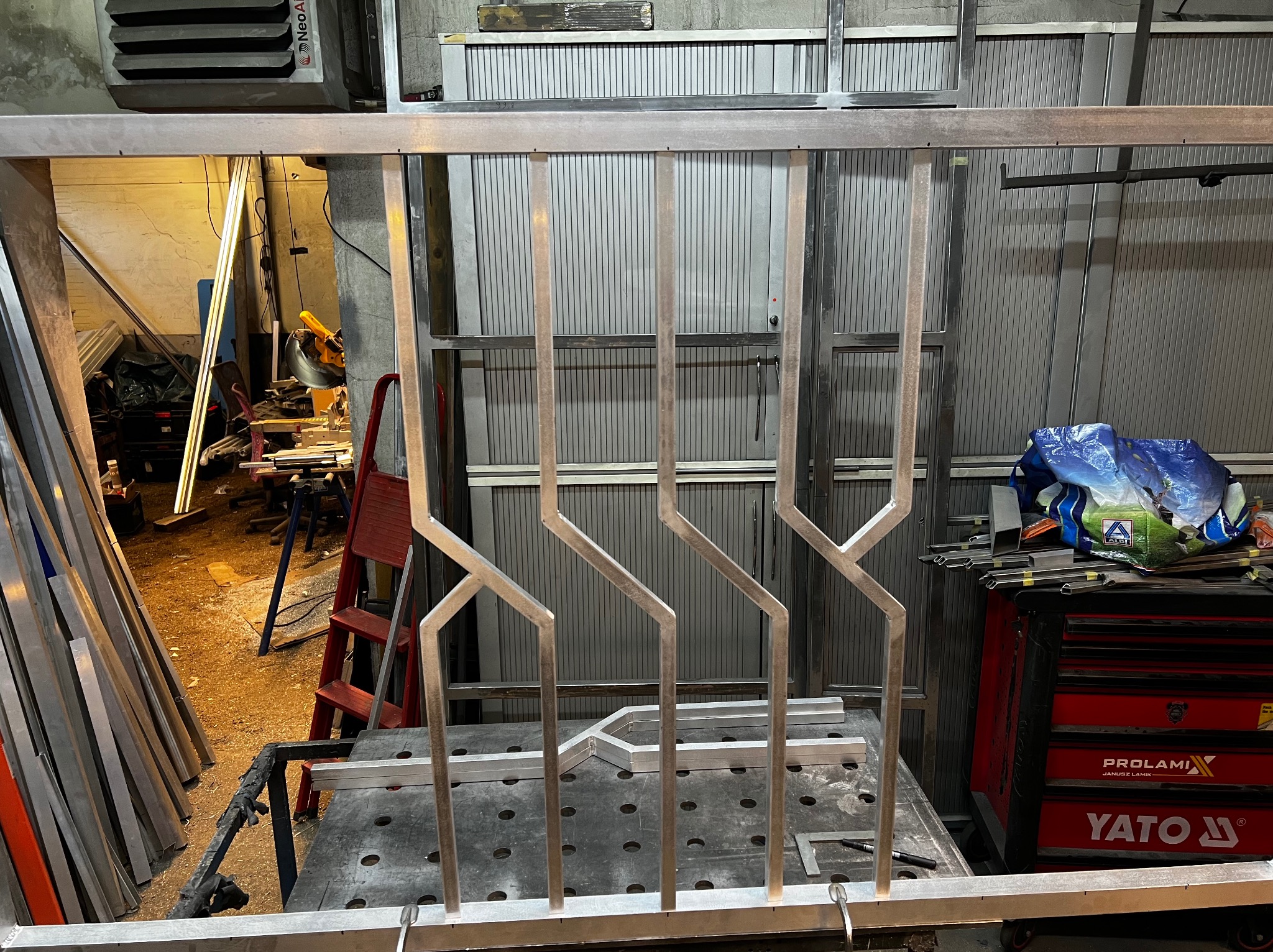 Metalowa balustrada w warsztacie, z geometrycznym wzorem. Widoczne narzędzia i materiały, co sugeruje etap produkcji. Balustrada wykonana z jasnego metalu.