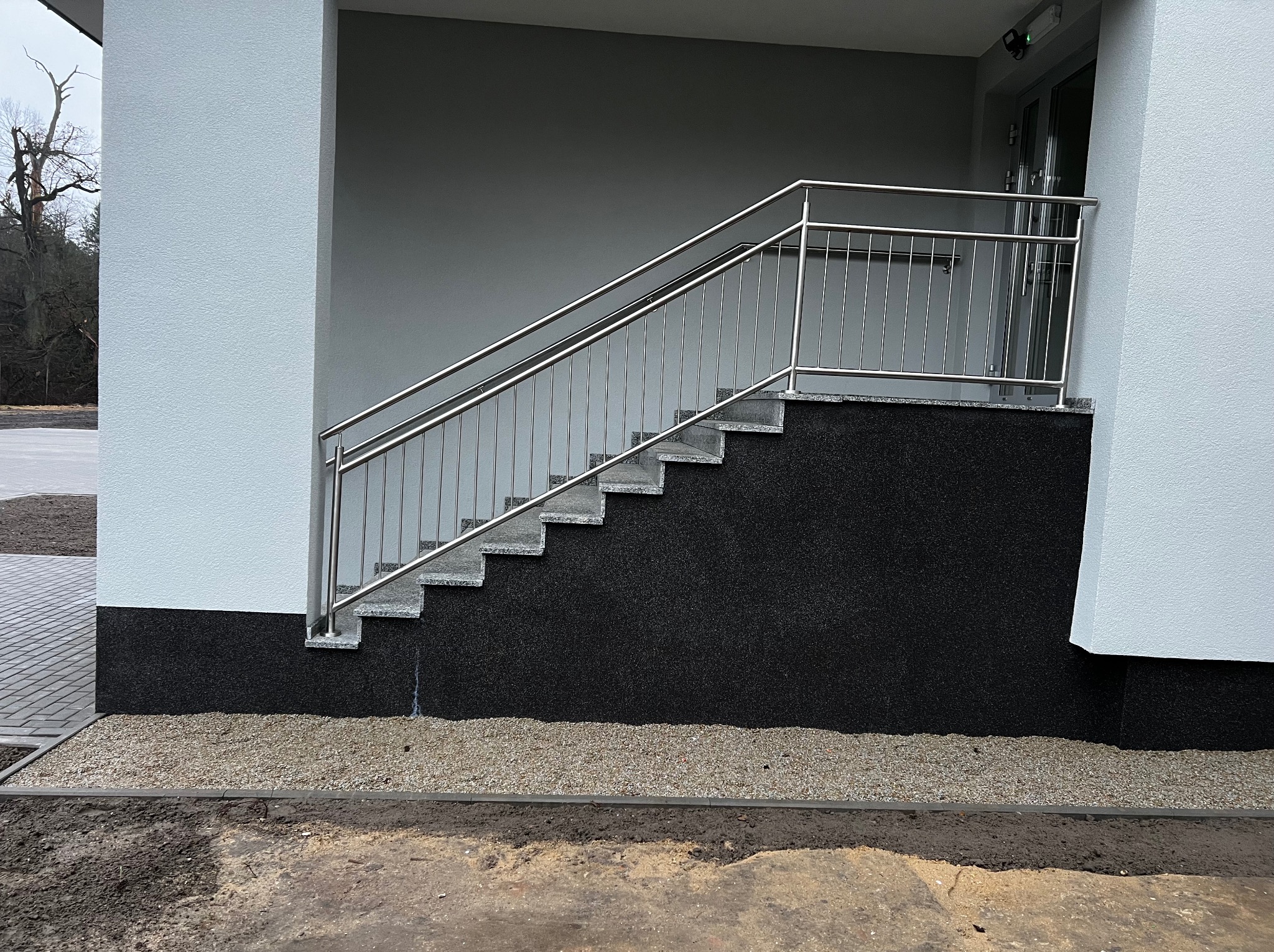 Zewnętrzne schody z szarymi stopniami i nowoczesną balustradą ze stali nierdzewnej. Tło stanowi elewacja budynku w kolorze popielatym. Widok z dołu.