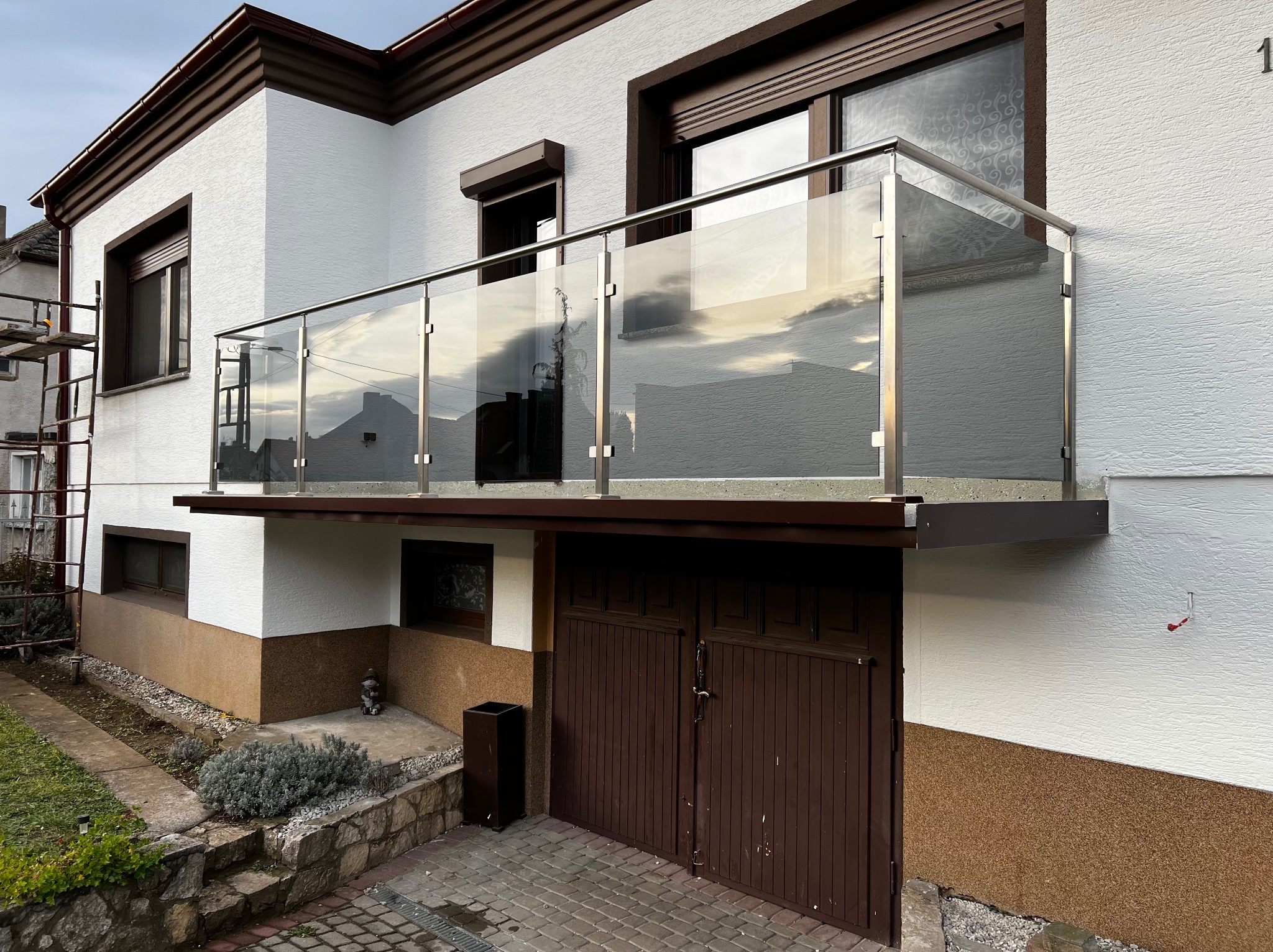 Szklana balustrada balkonowa z nierdzewnymi słupkami, zamontowana na budynku z garażem. Widoczne odbicia nieba i otoczenia w szkle.