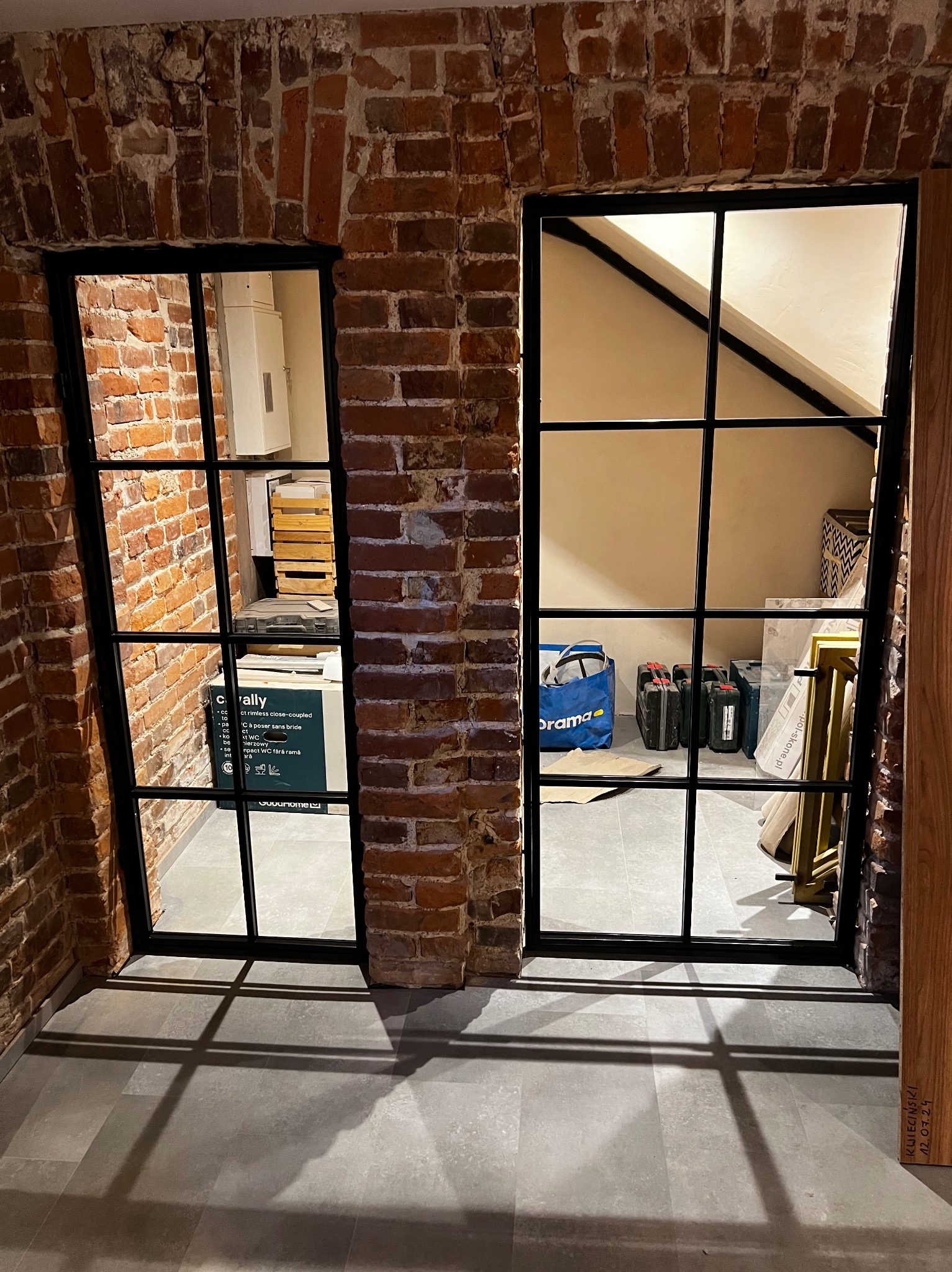 Dwa okna z czarnymi szprosami w murze z czerwonej cegły. W tle widoczne przedmioty w pomieszczeniu, w tym skrzynki i torba. Widoczne cienie na podłodze.