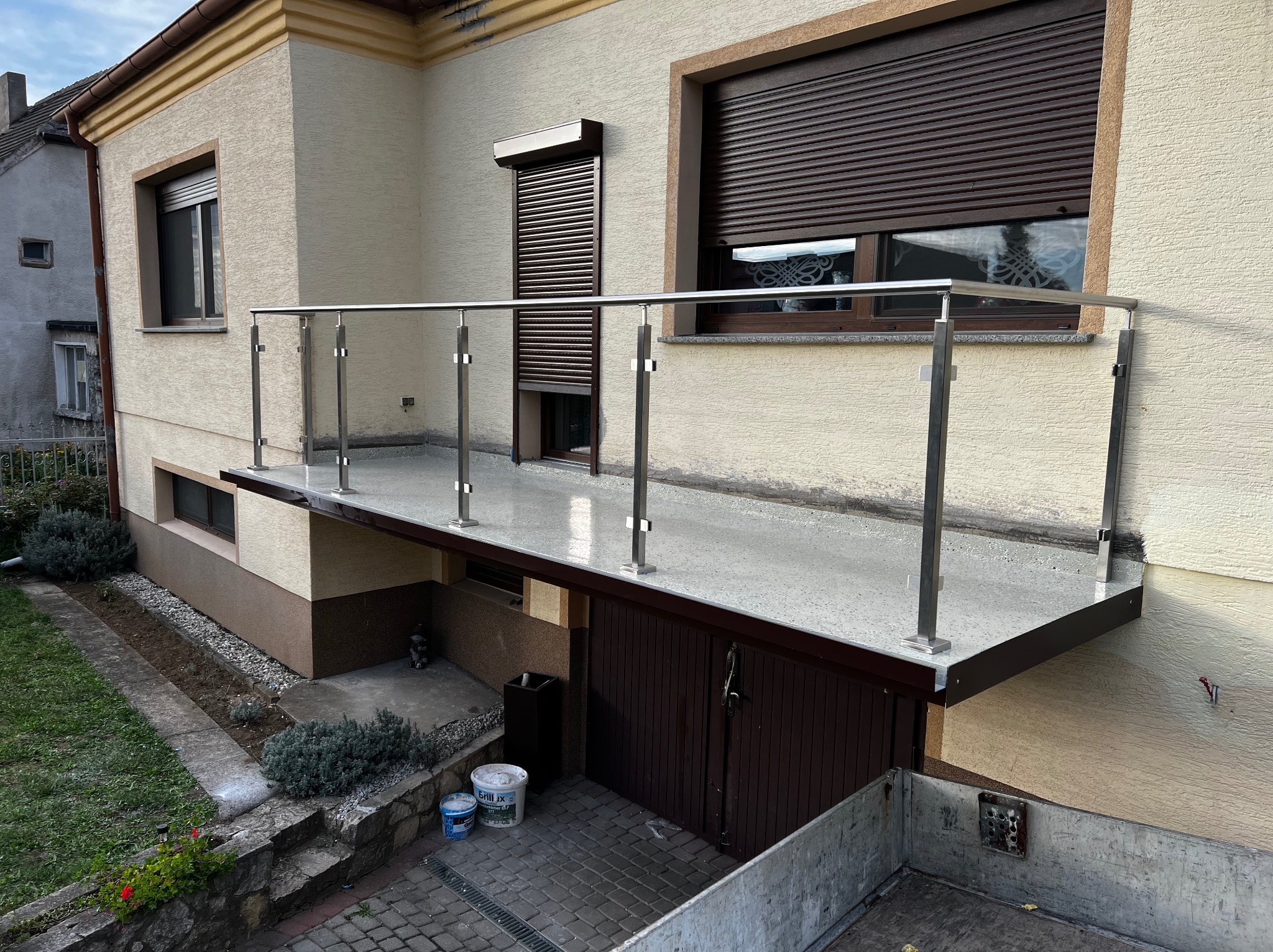 Nierdzewna balustrada balkonowa z pionowymi słupkami, zamontowana przy budynku z brązowymi roletami. Widok z dołu, z elementami ogrodu i wejścia do garażu.