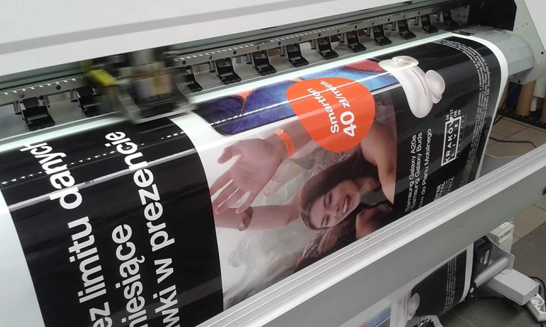Drukarka wielkoformatowa w trakcie drukowania plakatu reklamowego z wizerunkiem uśmiechniętej kobiety i informacją o ofercie smartfona w abonamencie.