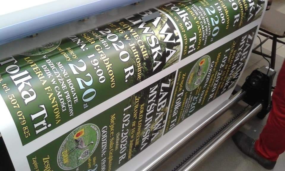 Drukarka wielkoformatowa w trakcie drukowania banerów reklamowych z motywem myśliwskim i informacjami o zabawie, widoczne fragmenty rolek z nadrukowanymi plakatami.