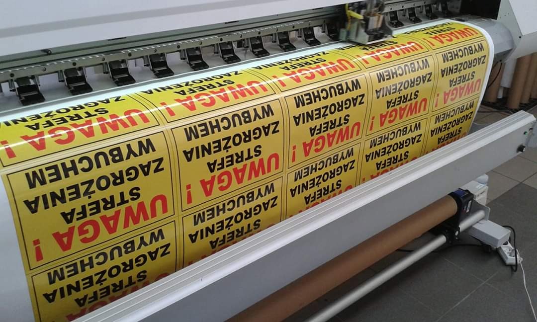 Drukarka wielkoformatowa drukująca rolkę żółtych naklejek ostrzegawczych z napisem 'UWAGA! STREFA ZAGROŻENIA WYBUCHEM'
