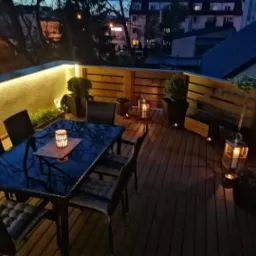 Oświetlony taras z drewnianą podłogą i balustradą, stolik z czarnym, błyszczącym blatem i plecione krzesła z poduszkami, wieczorne oświetlenie LED i lampiony ze świecami, widok na budynki w tle.