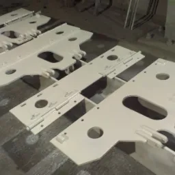 Białe, pomalowane elementy konstrukcji stalowej z otworami i zawiasami, leżące na podłodze w hali produkcyjnej.