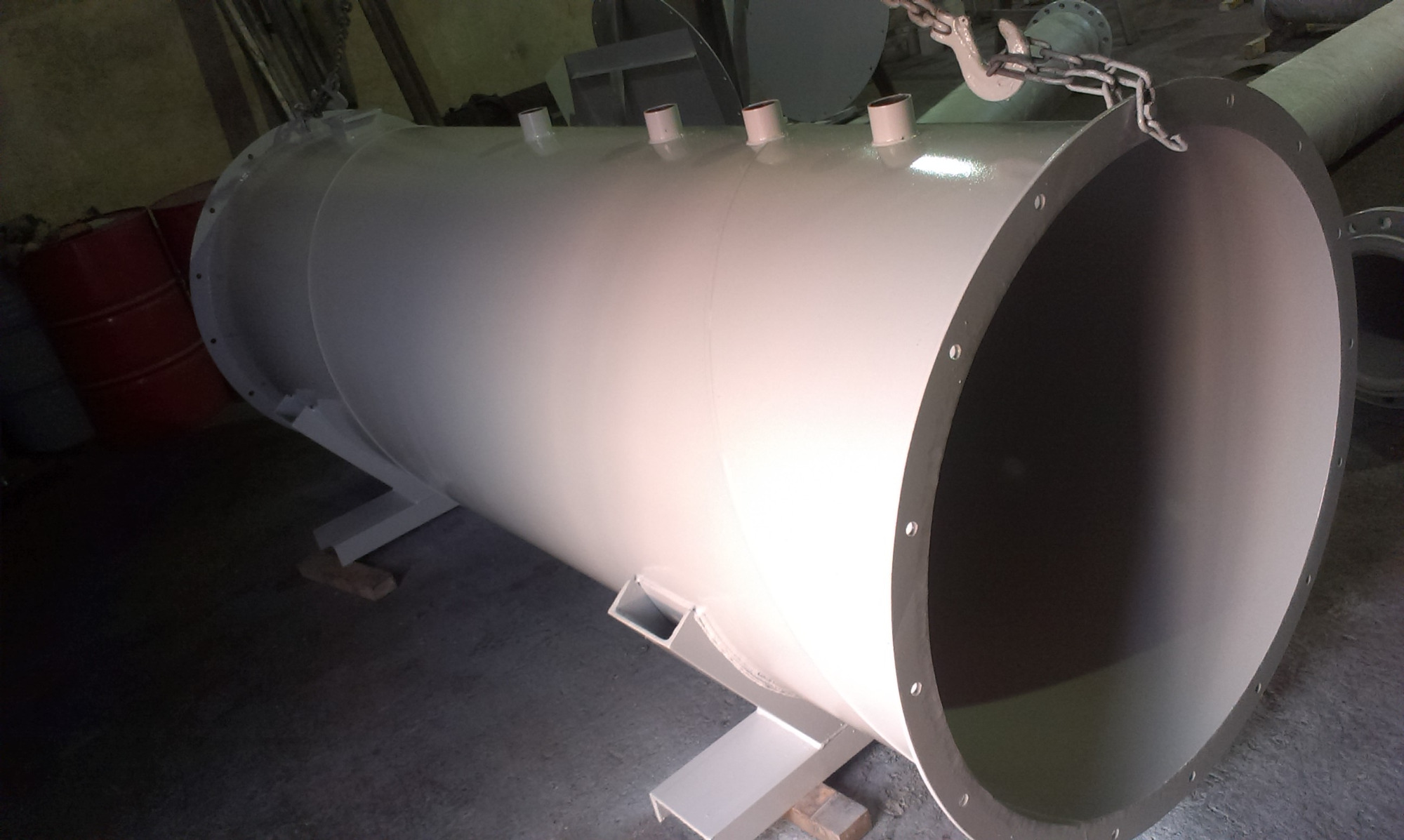 Malowany na jasnoszaro cylindryczny zbiornik stalowy z króćcami, podparty na wspornikach, transportowany za pomocą łańcucha.