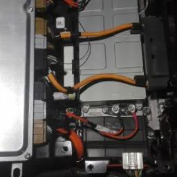 Widok z góry na akumulator hybrydowy w komorze silnika, widoczne pomarańczowe kable wysokiego napięcia i złącza elektryczne.