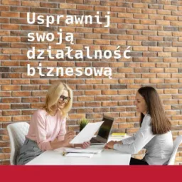 Dwie kobiety przy biurku omawiają dokumenty, laptop w tle, ceglana ściana z napisem 'Usprawnij swoją działalność biznesową' w tle.