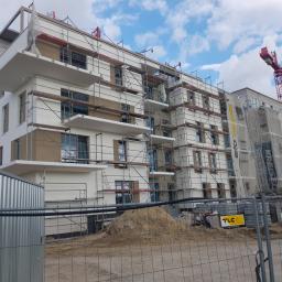 Mrówex - Budynek wielorodzinny w trakcie prac elewacyjnych, widoczne rusztowania, częściowo otynkowane i pomalowane fragmenty ścian, w tle żuraw budowlany i błękitne niebo z chmurami.