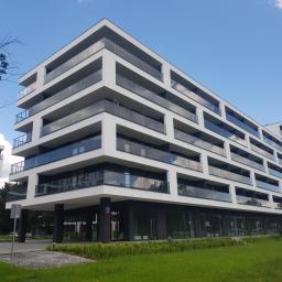 Mrówex - Nowoczesny budynek apartamentowy z białymi elewacjami i szklanymi balkonami, widok z poziomu trawnika w słoneczny dzień.