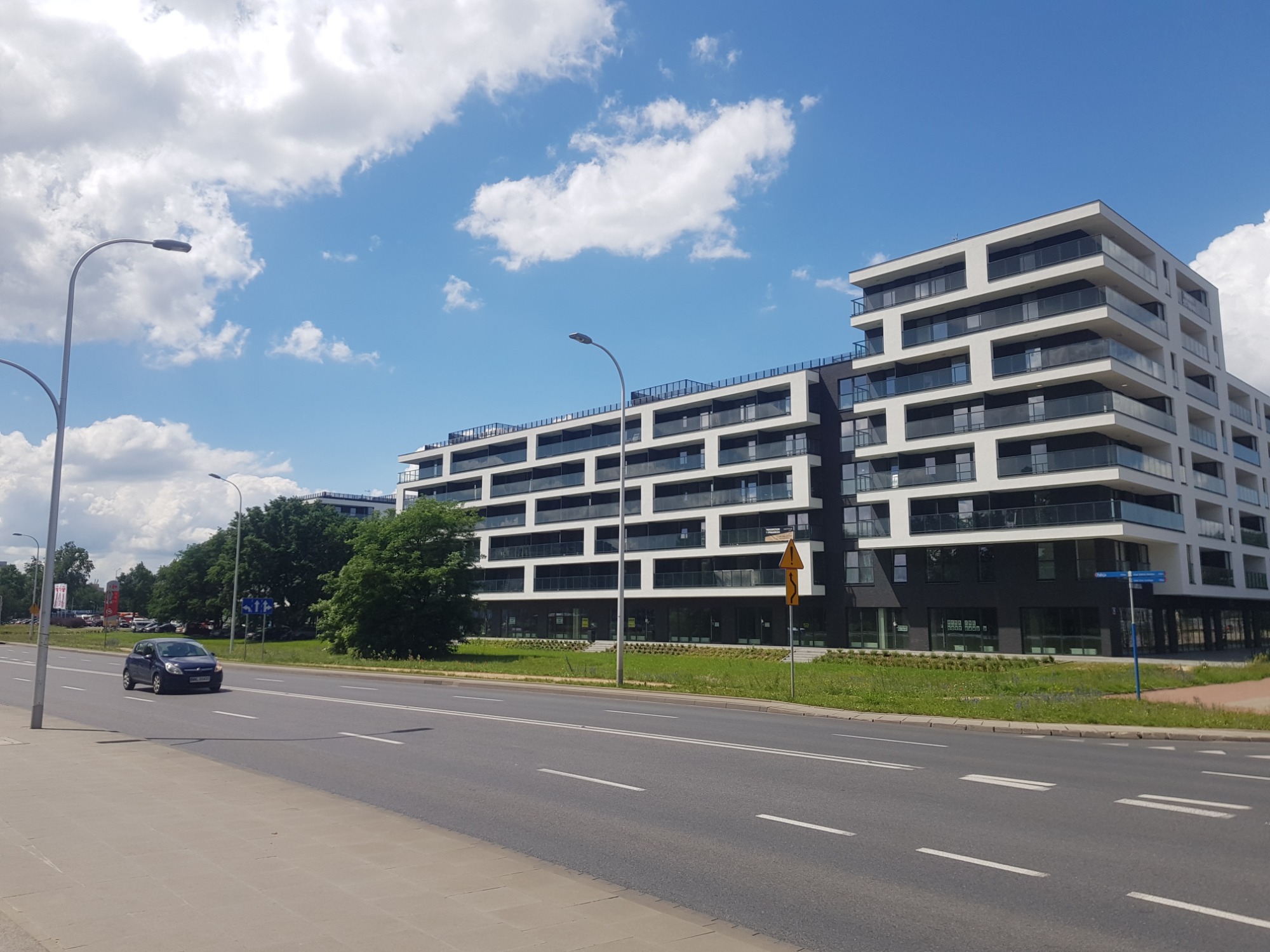 Nowoczesny budynek apartamentowy z białymi elewacjami i czarnymi akcentami, widoczny z perspektywy ulicy z przejeżdżającym samochodem i błękitnym niebem w tle.