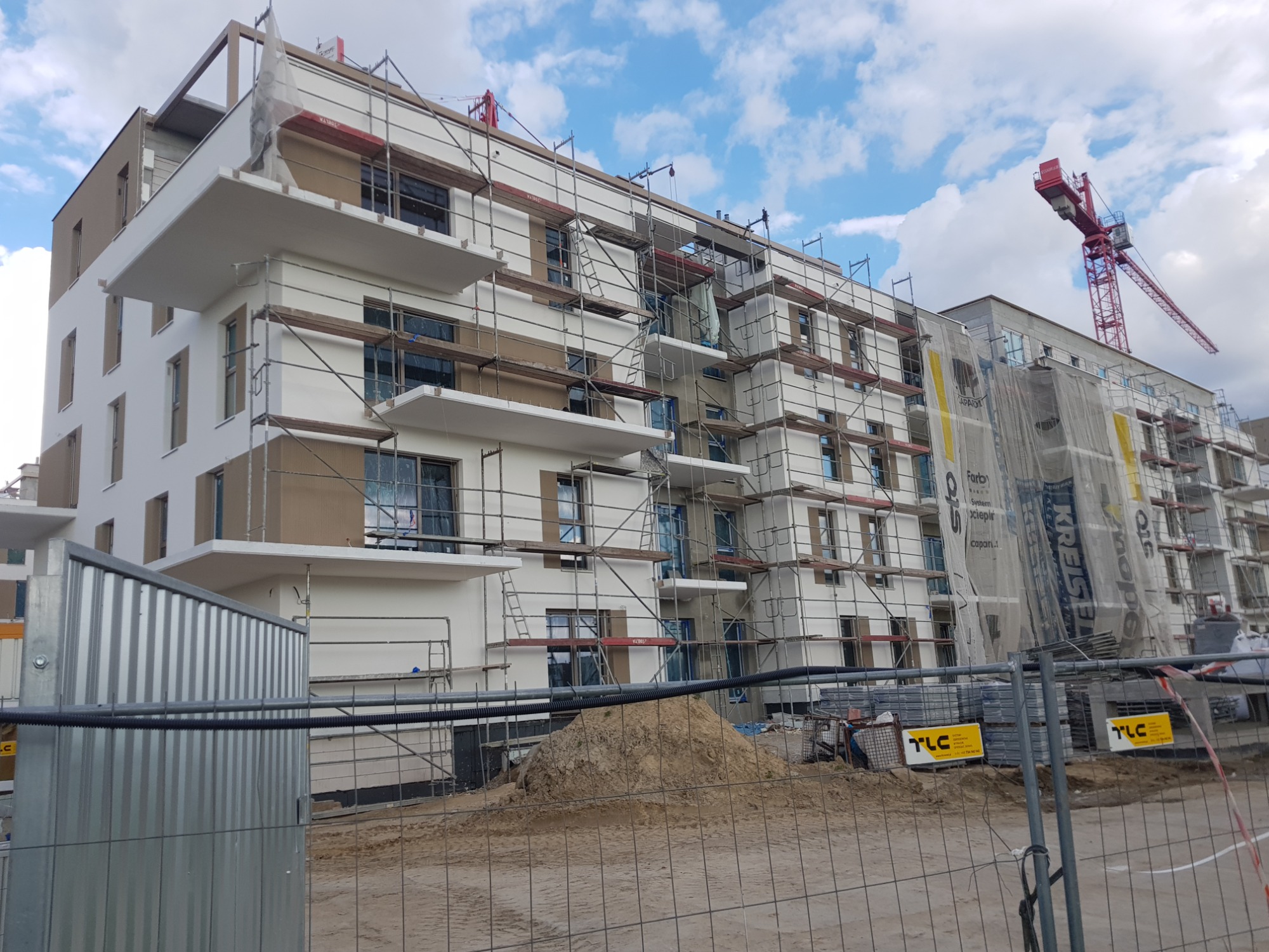 Budynek wielorodzinny w trakcie prac elewacyjnych, widoczne rusztowania, częściowo otynkowane i pomalowane fragmenty ścian, w tle żuraw budowlany i błękitne niebo z chmurami.