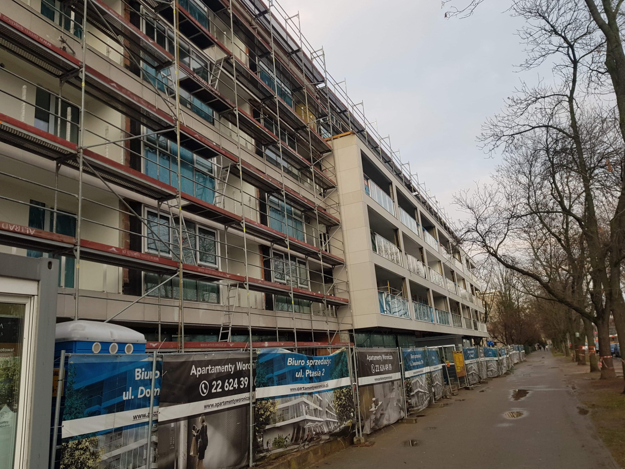 Wielopiętrowy budynek w trakcie prac elewacyjnych, otoczony rusztowaniami i tymczasowym ogrodzeniem z reklamami biura sprzedaży mieszkań. Widoczna przenośna toaleta.