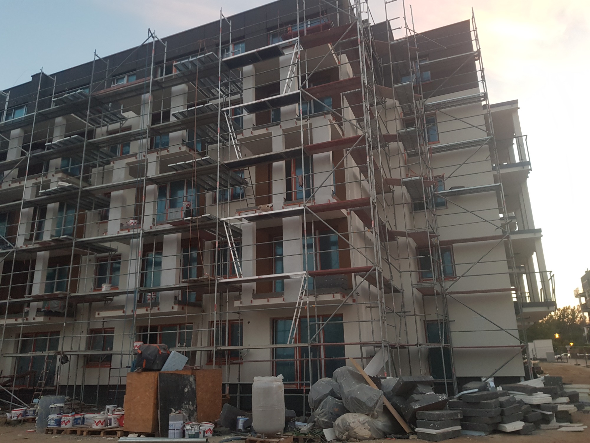 Wielopiętrowy budynek w trakcie prac elewacyjnych, otoczony rusztowaniami, z widocznymi materiałami budowlanymi i farbami w tle, uchwycony pod kątem z dołu.