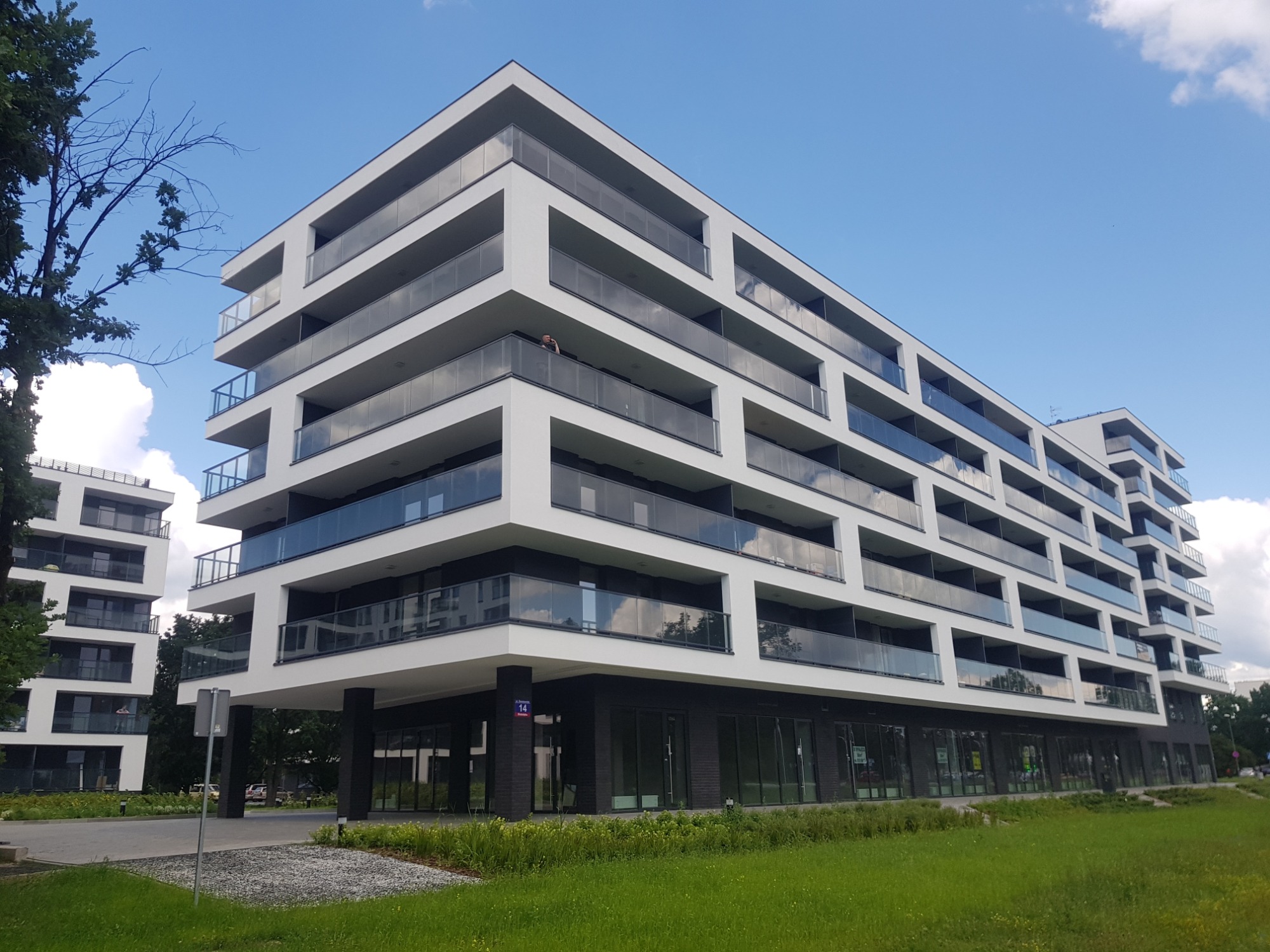Nowoczesny budynek apartamentowy z białymi elewacjami i szklanymi balkonami, widok z poziomu trawnika w słoneczny dzień.