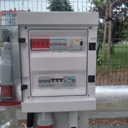 Otwarta skrzynka elektryczna z widocznymi bezpiecznikami i wyłącznikami, zamontowana na zewnątrz budynku, z widocznymi gniazdami i przewodami, tło stanowi metalowe ogrodzenie i zieleń.