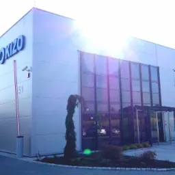 Nowoczesna hala przemysłowa z szarej blachy trapezowej, z niebieskim logo 'OKIZO' na fasadzie, widoczne okna, brama wjazdowa i szlaban, oświetlona wschodzącym słońcem.