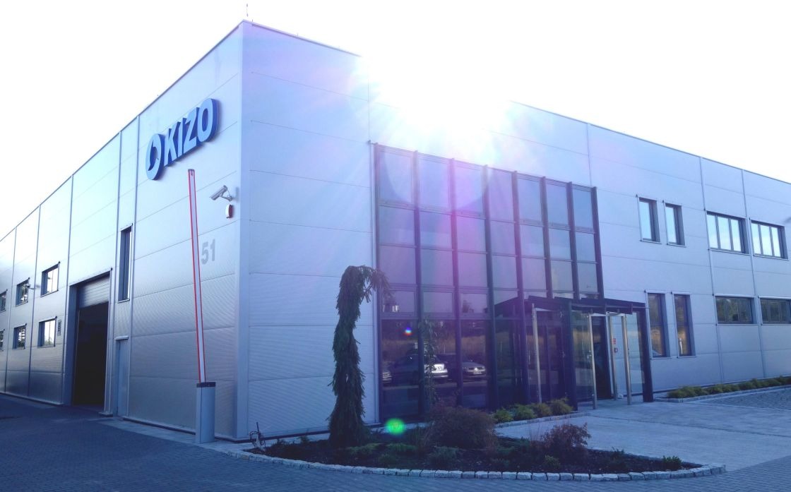 Nowoczesna hala przemysłowa z szarej blachy trapezowej, z niebieskim logo 'OKIZO' na fasadzie, widoczne okna, brama wjazdowa i szlaban, oświetlona wschodzącym słońcem.
