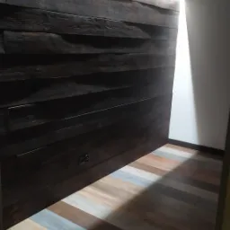 Ciemna, rustykalna ściana z desek z ukrytymi gniazdkami elektrycznymi, kontrastująca z podłogą ułożoną z paneli imitujących drewno w różnych odcieniach, oświetlona bocznym światłem.