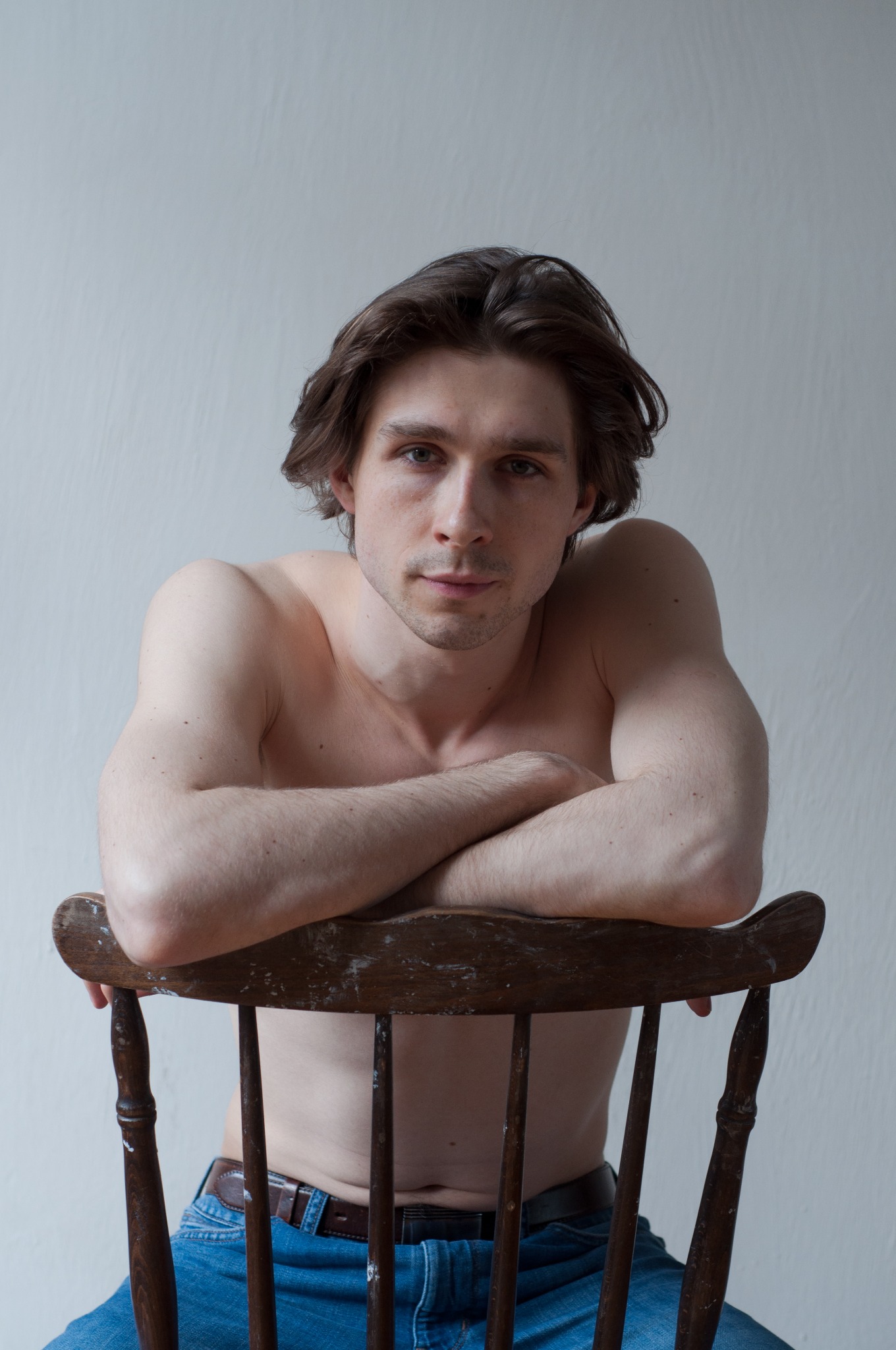 Portret młodego mężczyzny bez koszuli, opierającego się o stare, drewniane krzesło z widocznymi śladami użytkowania, na tle białej ściany.