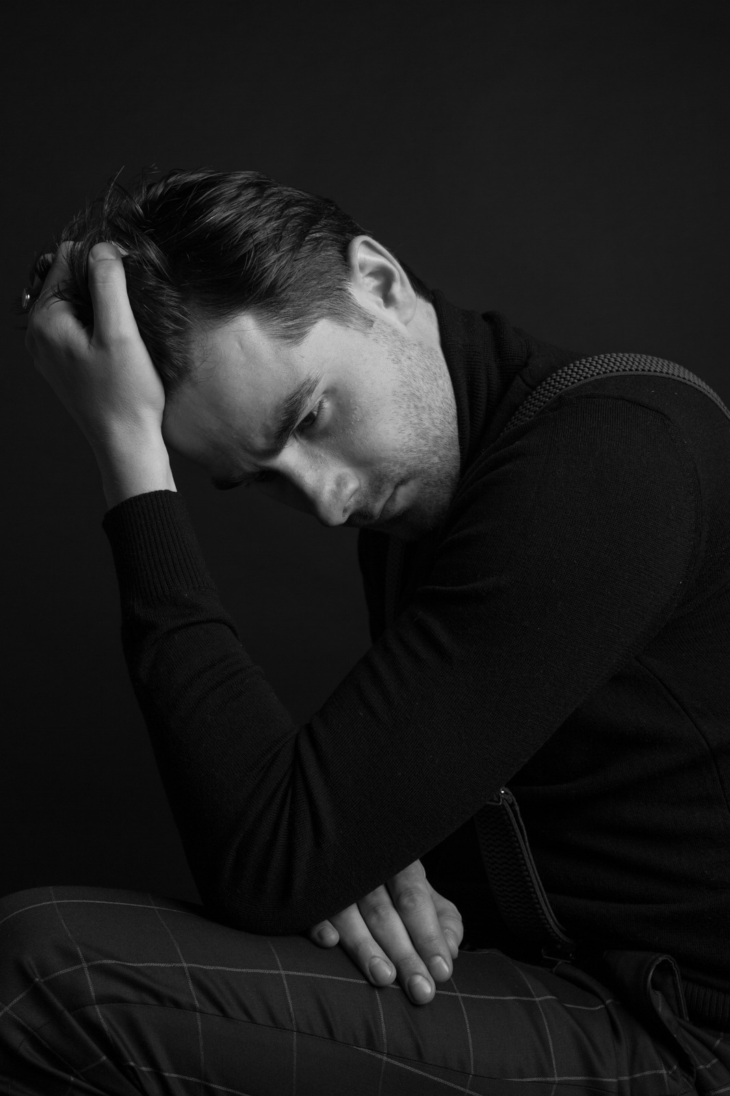 Czarno-biały portret mężczyzny w ciemnym swetrze, z głową opartą na dłoni, wzrok skierowany w dół, na ciemnym tle.
