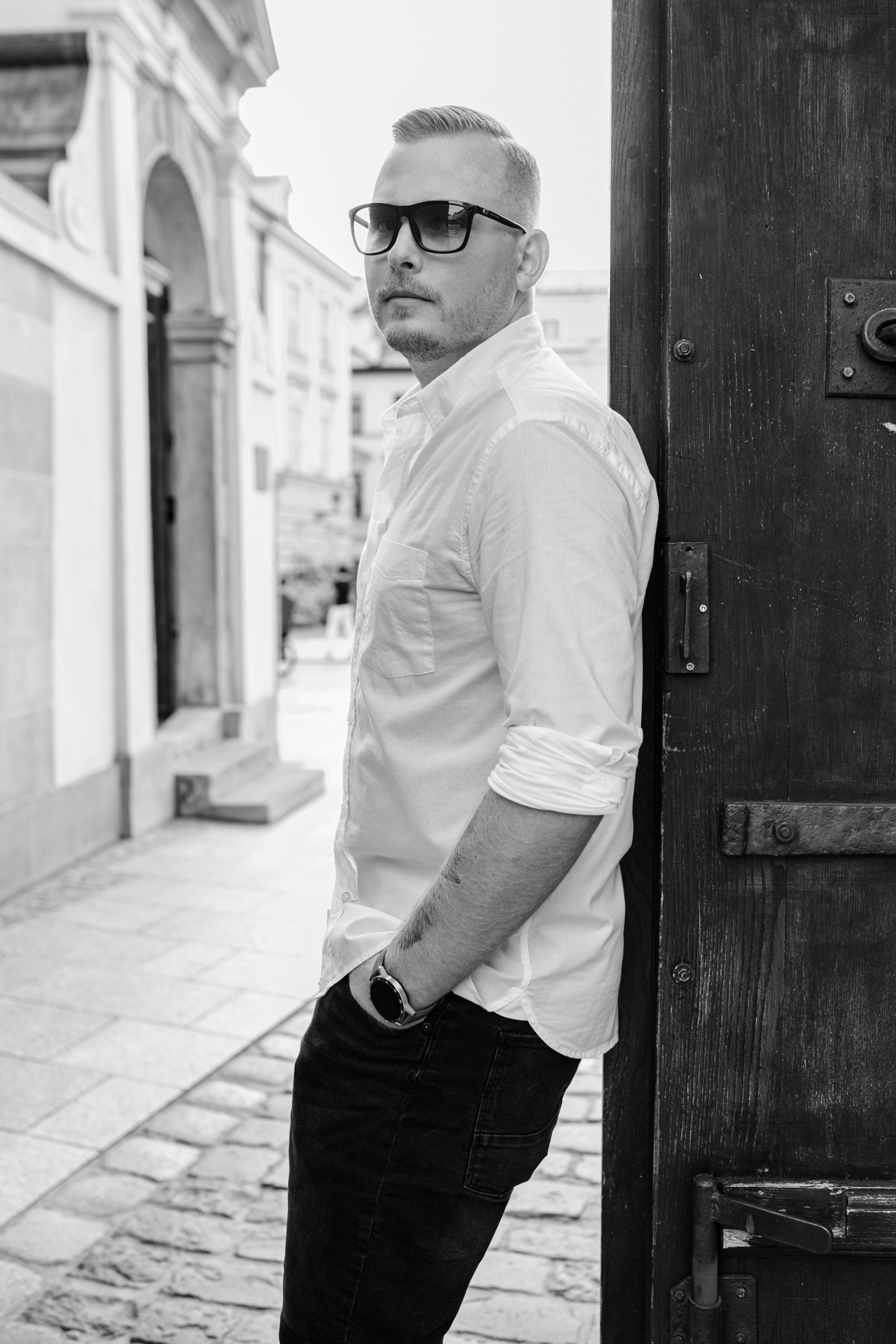 Mężczyzna w okularach przeciwsłonecznych, oparty o stare drewniane drzwi na tle kamiennej uliczki w czarno-białej fotografii.