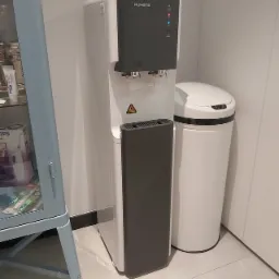 Nowoczesny, biało-szary dystrybutor wody RUKENS z wyświetlaczem LED, umieszczony w rogu pomieszczenia obok białego kosza na śmieci z czujnikiem ruchu, widoczny fragment przeszklonej szafki...