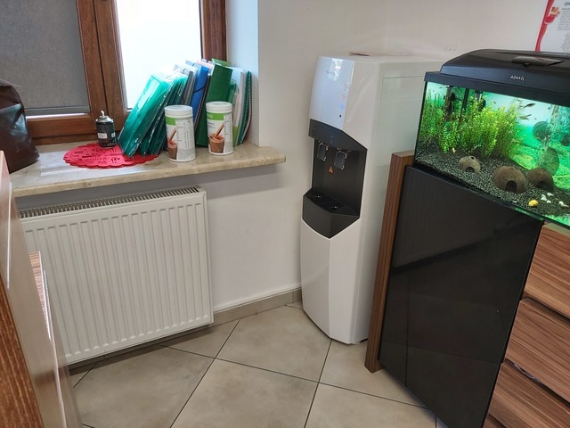 Biały dystrybutor wody w biurze, obok akwarium z rybkami i szafka, na parapecie okna segregatory i puszki z suplementami diety