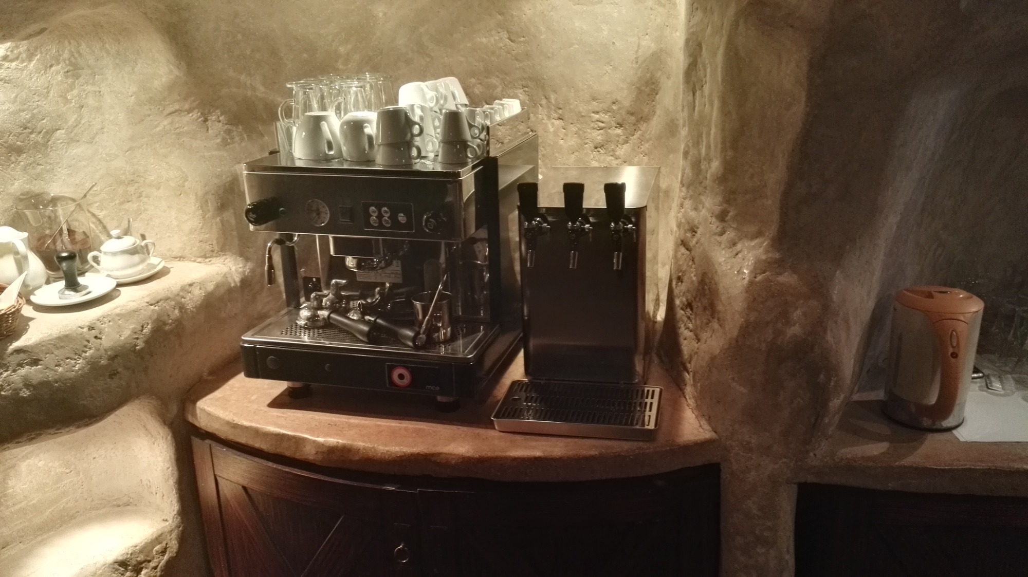 Profesjonalny ekspres do kawy z dystrybutorem wody i stosem filiżanek na blacie w kamiennym wnętrzu.