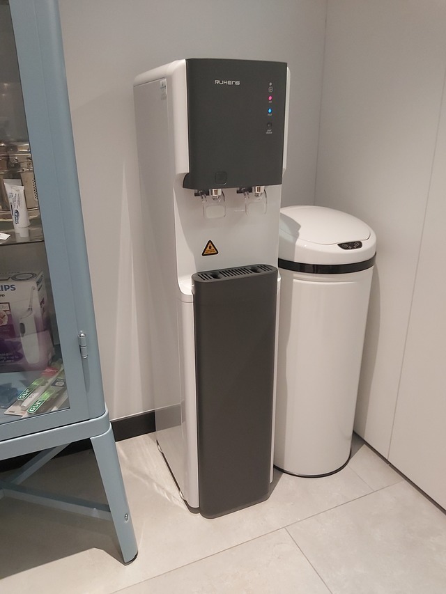 Nowoczesny, biało-szary dystrybutor wody RUKENS z wyświetlaczem LED, umieszczony w rogu pomieszczenia obok białego kosza na śmieci z czujnikiem ruchu, widoczny fragment przeszklonej szafki...