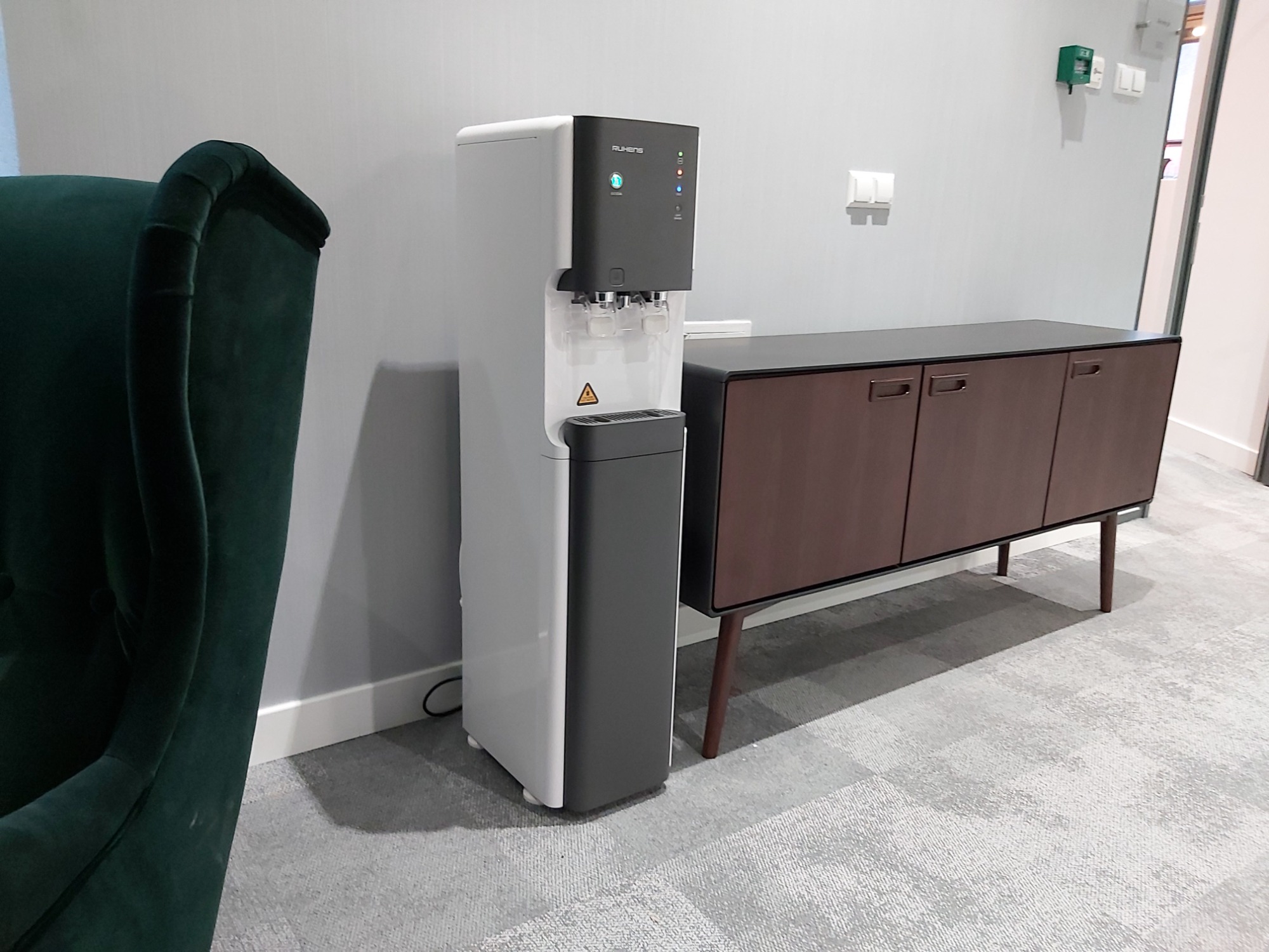 Nowoczesny dystrybutor wody Ruhens w biurowym wnętrzu, obok ciemnozielonego fotela i brązowej komody na nóżkach.