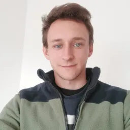 Portret młodego mężczyzny z niebieskimi oczami w zielonej polarowej kurtce na tle białej ściany.