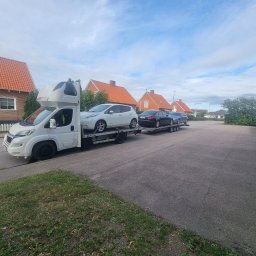 RAF-TRANS Rafał Matwiejczyk - Biała laweta z kabiną sypialną przewozi trzy samochody osobowe, w tym biały samochód elektryczny Nissan Leaf na górnym poziomie, na tle zabudowy mieszkalnej z czerwonymi dachami.
