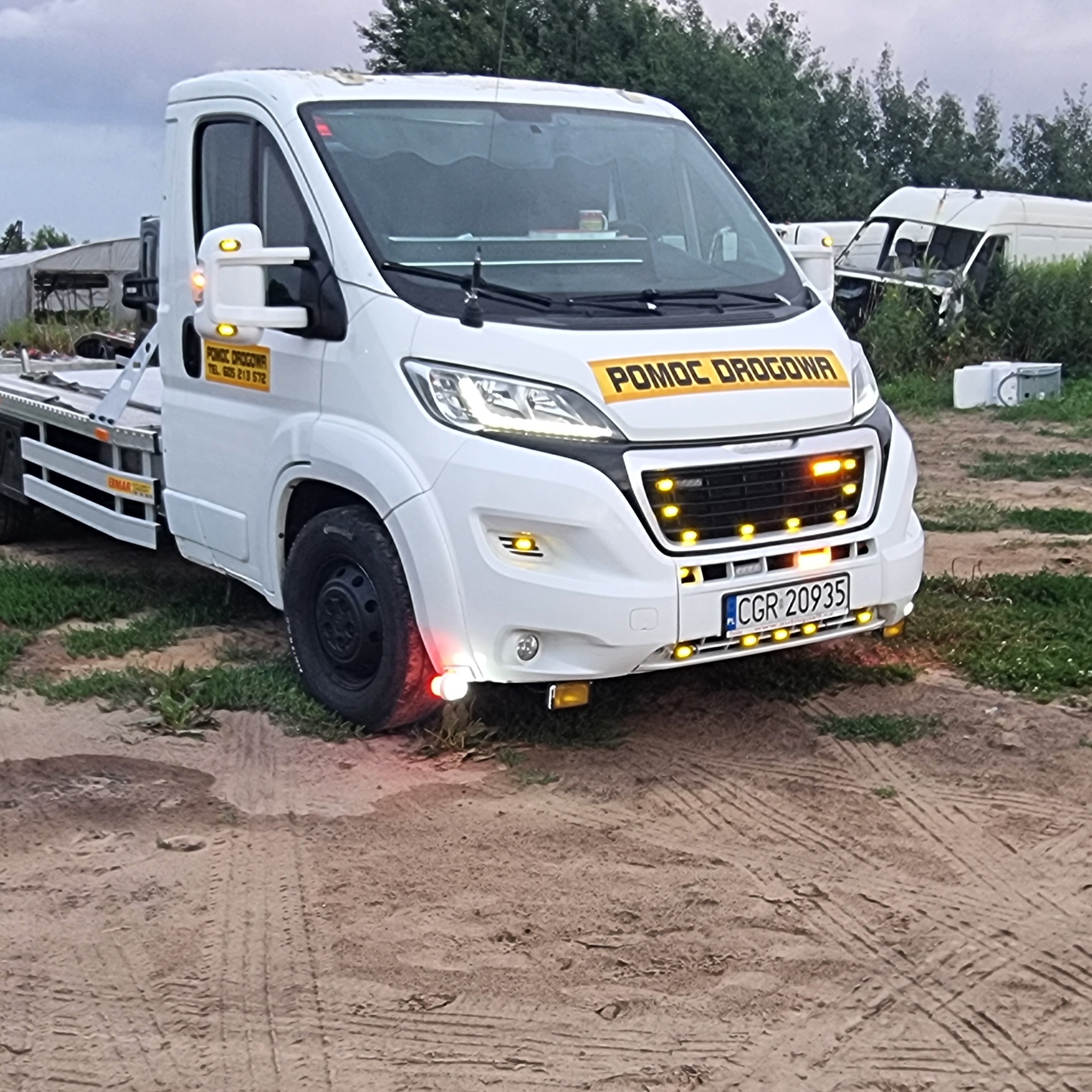 Biała laweta z napisem 'Pomoc Drogowa' i włączonymi światłami awaryjnymi, zaparkowana na piaszczystym terenie z widocznymi śladami opon, w tle zarośla i uszkodzony pojazd.