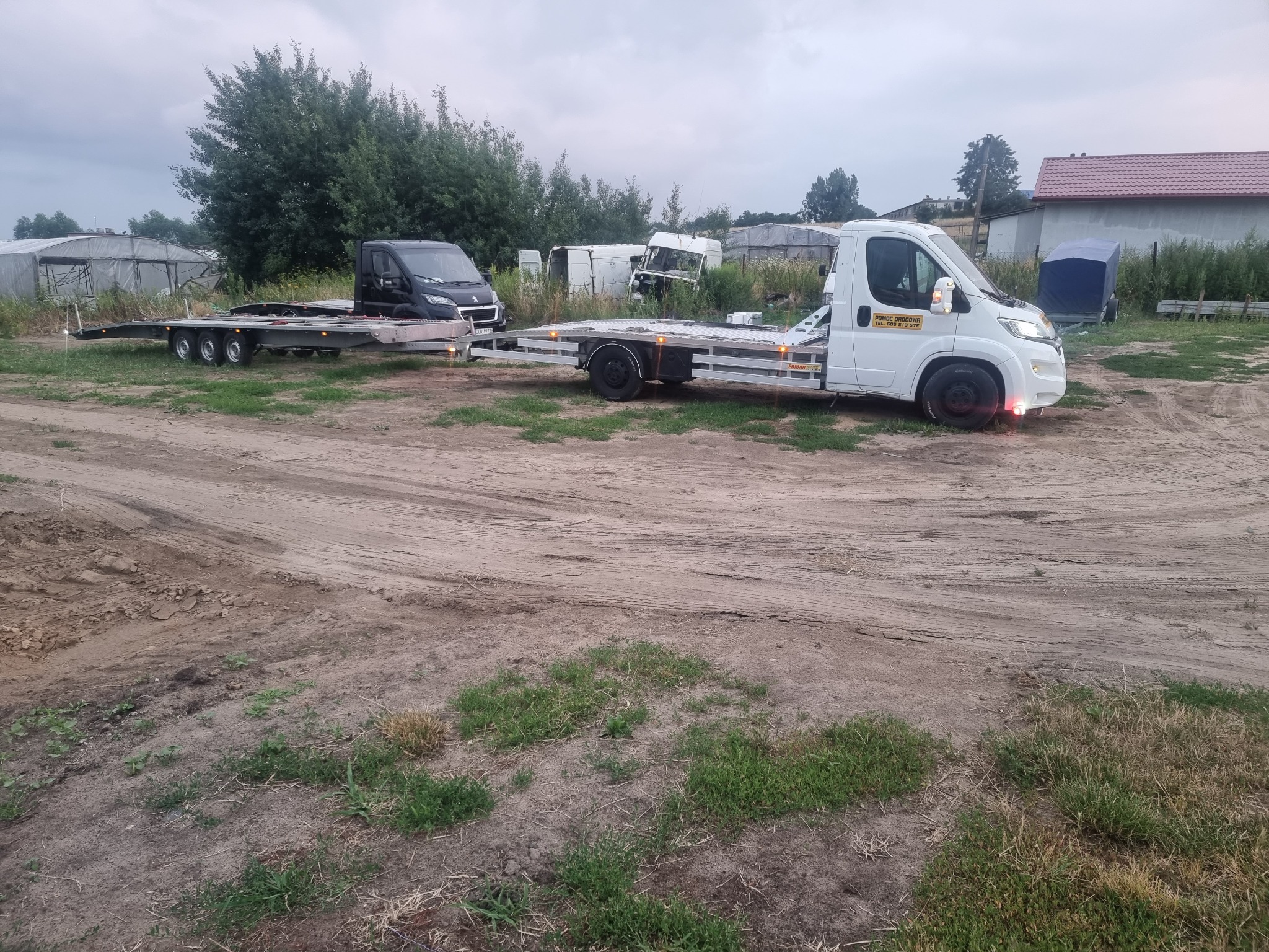 Czarno-biały zestaw lawety i przyczepy na piaszczystym terenie, z zaroślami i budynkami w tle, sfotografowany w pochmurny dzień.