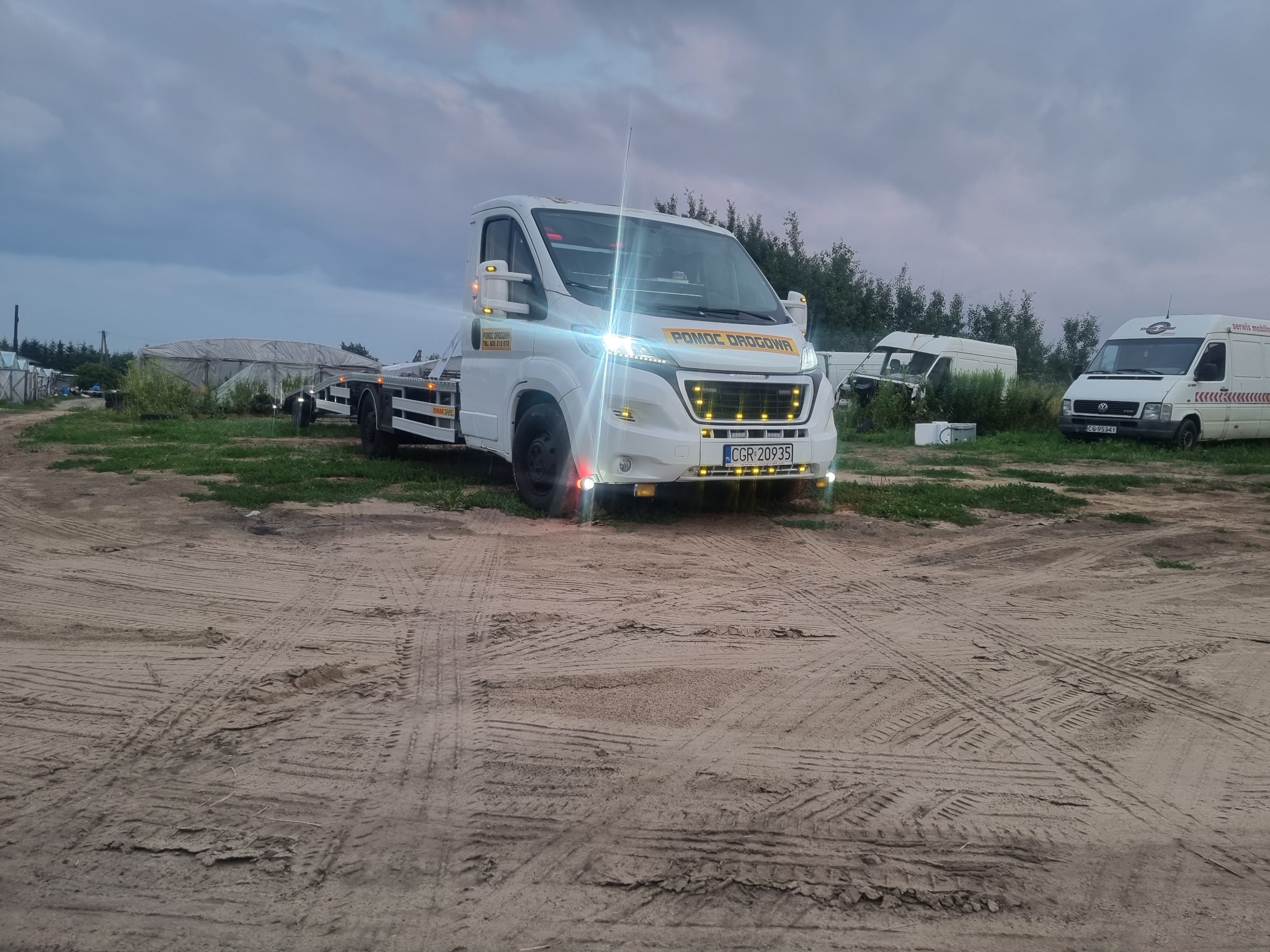Biała laweta z napisem 'POMOC DROGOWA' na tle piaszczystego terenu z widocznymi śladami opon i innymi pojazdami w tle, sfotografowana o zmierzchu.