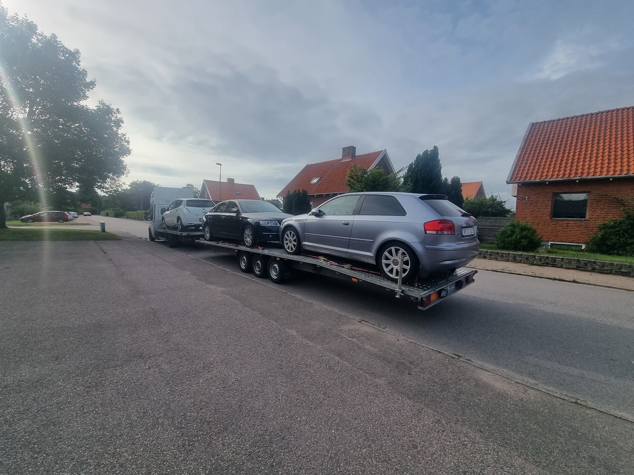 Trzy samochody osobowe, srebrny, czarny i biały, zabezpieczone na platformie lawety, zaparkowane na ulicy w zabudowie jednorodzinnej, widok z boku.