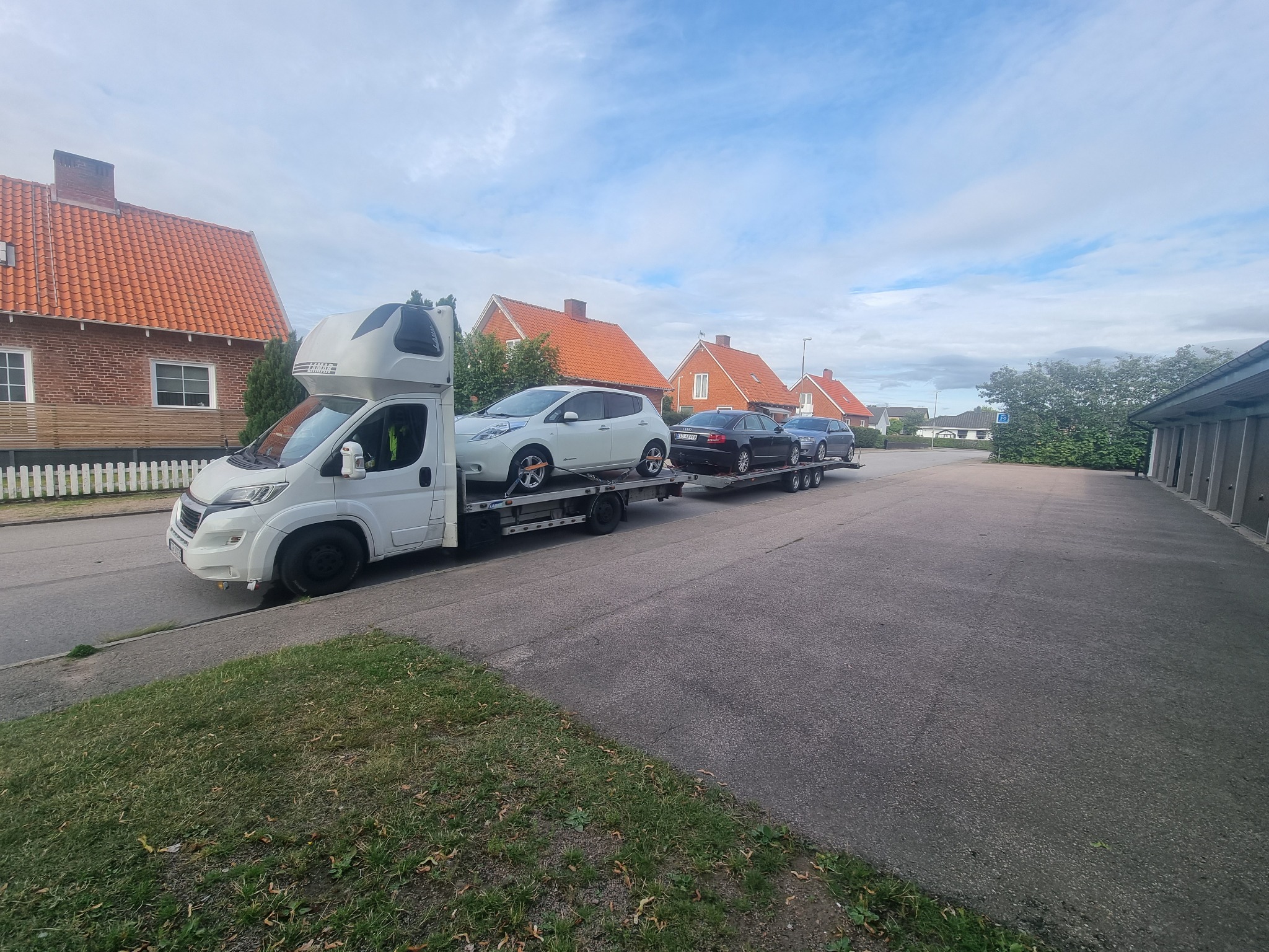 Biała laweta z kabiną sypialną przewozi trzy samochody osobowe, w tym biały samochód elektryczny Nissan Leaf na górnym poziomie, na tle zabudowy mieszkalnej z czerwonymi dachami.